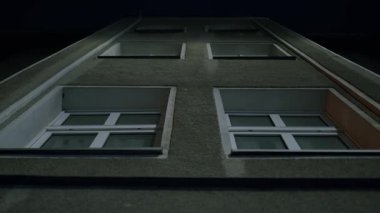 Gece bir apartmanın alçak açılı görüntüsü. Pencerelerin tekrar eden şekli geometrik bir bileşim oluşturuyor. Karanlık gökyüzü, mimarinin katıksız, kentsel hissini arttırır..