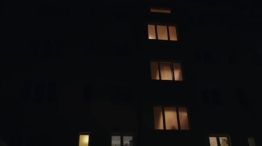 Bazı pencereleri aydınlatılmış bir apartmanın gece görüşü bir ışık ve gölge deseni yaratıyor. Geceleri kentsel yaşam ve konut mimarisini temsil eder..