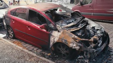 Kırmızı bir araba, yangından ağır hasar görmüş, ön yarısı tamamen tahrip olmuş. Yanmış ve el değmemiş kısımlar arasındaki zıtlık ateşin yıkıcı gücünü vurguluyor. Şehir kazası sahnesi.