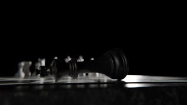 Düşmüş siyah satranç kralı dramatik bir yakın plan çekimde satranç tahtasında yatıyor. Yenilgiyi ve stratejik savaşın sonucunu sembolize ediyor. Beyaz parçalar bulanık arkaplanda muzaffer duruyor.