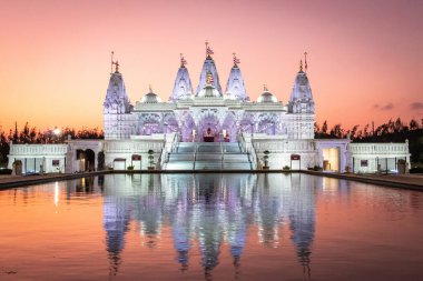 Purple illuminated BAPS Shri Swaminarayan Mandir reflected against water during a purple sunset. Photo taken in Houston Texas clipart