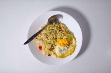 Beyaz bir kase sebzelerle karıştırılmış bir pilav, yanında sahanda yumurta ve tabakta dinlenen minimum ışık arka planında sergilenen bir kaşık içerir..