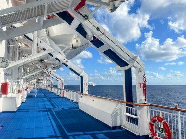 Mavi yolu, filikaları ve açık gökyüzünün altındaki okyanusu olan bir yolcu gemisinin panoramik görüntüsü. Güvertenin yapısı ve deniz manzarasının temiz çizgileri bir seyahat ve rahatlama atmosferi yaratıyor..