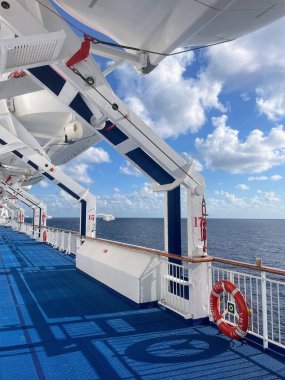 Mavi yolu, filikaları ve açık gökyüzünün altındaki okyanusu olan bir yolcu gemisinin panoramik görüntüsü. Güvertenin yapısı ve deniz manzarasının temiz çizgileri bir seyahat ve rahatlama atmosferi yaratıyor..