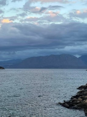 Sakin suların, kayalık kıyı şeridinin ve dramatik bulutlu bir gökyüzünün altındaki uzak dağların bulunduğu sakin bir kıyı manzarası. Huzurlu atmosfer, huzurlu bir deniz kıyısındaki doğanın güzelliğini yansıtıyor.