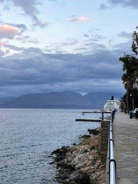 Dramatik bulutlu bir gökyüzünün altında rıhtıma yanaşmış bir yolcu gemisiyle manzaralı bir sahil geçidi. Sakin deniz, sakin bir akşam atmosferini yansıtıyor. Uzaktaki dağlar görülebiliyor.