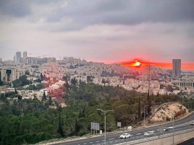 İsrail, Kudüs 'te eşsiz bir gün batımı. Şehrin üzerinde parlayan kırmızı bir güneş var. Canlı renkler ve dramatik ışık büyüleyici bir manzara yaratır, kentsel ve doğal güzellikleri harmanlar.