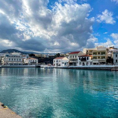 Yunanistan 'ın Girit adasında sakin turkuaz su, geleneksel binalar ve tüylü bulutlarla dolu dramatik bir gökyüzünün yer aldığı güzel bir liman manzarası. Sakin atmosfer bu Akdeniz kıyı kasabasının cazibesini yansıtıyor.