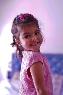 Fotoğraf için poz verirken kameraya gülümseyen pembe elbiseli bronzlaşmış çocuk.