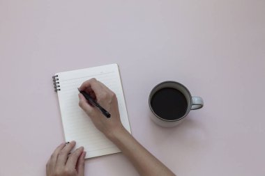 Pembe arka planda, bir fincan sade kahvenin yanında, defterin üzerine kadın el yazısının üst görüntüsü. Boşluğu kopyala