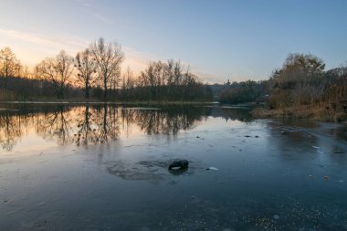 Kış günbatımında gölde ağaçlar ön planda.