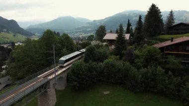 Alplerdeki küçük Gstaad kasabasından geçen bir trenin destansı hava aracı görüntüleri. Chalet ve etrafındaki güzel dağlar. Yüksek kalite 4k görüntü