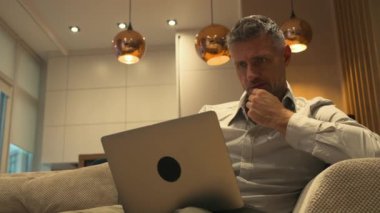 Gri saçlı yakışıklı bir adam evde dizüstü bilgisayarıyla oturuyor iş kıyafeti içinde ve ticaret yapıyor. Yüksek kalite 4k görüntü