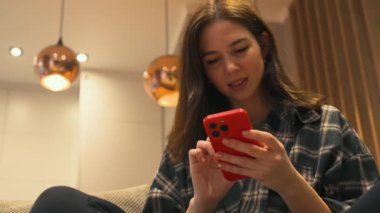 Güzel, genç, esmer kadın oturma odasındaki kanepede oturuyor. Telefonu ellerinde ve gülümsüyor. Ekrandaki bir şeyi kaydırmak ve keyif almak. - Evet. Yüksek kalite 4k görüntü
