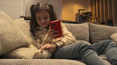 7-8 yaşlarında rahat giyimli tatlı bir kız, bir kanepede oturuyor, telefonuna odaklanmış, ve hafifçe beyaz faresini okşuyor. Yüksek kalite 4k görüntü