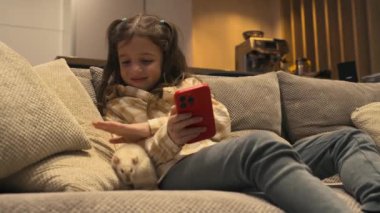 7-8 yaşlarında rahat giyimli tatlı bir kız evdeki bir kanepede oturuyor, telefonla oynuyor ve beyaz faresini okşuyor. Yüksek kalite 4k görüntü