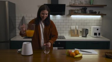 Gülümseyen güzel esmer kadın günlük kıyafetleriyle bardağa taze meyve suyu dolduruyor ve evdeki modern bir mutfakta içiyor. Yüksek kalite 4k görüntü