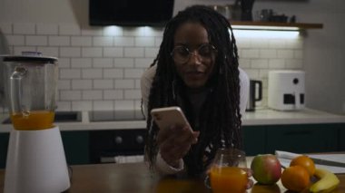 Modern mutfaktaki saç örgülü, gözlüklü, elinde telefon ve portakal suyu olan Afrikalı bir kadının portresi. Yüksek kalite 4k görüntü