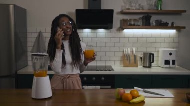 Uzun örgülü ve gözlüklü Afrikalı genç bir kadın modern bir mutfakta portakal suyu içerken telefonla konuşuyor. Mekanın samimi ve çağdaş bir havası var. Yüksek kalite 4k görüntü