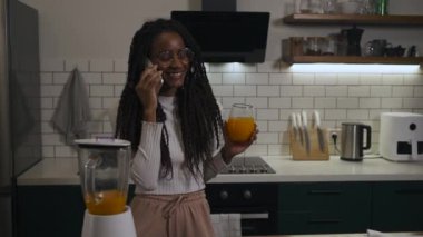 Amerikan kadın portresi, uzun örgülü ve gözlüklü modern bir mutfakta portakal suyu içerken telefonla konuşuyor. Yüksek kalite 4k görüntü