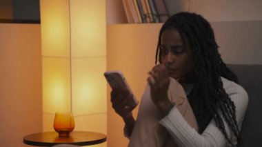 Güzel Afrikalı bir kadının, günlük kıyafetleriyle oturma odasındaki bir kanepede rahatlarken ve kaydırırken yakın plan çekimleri. Yüksek kalite 4k görüntü