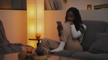 Saçları örgülü Afrikalı Amerikalı bir kadın rahat bir odada kanepede oturmuş telefonuna bakıyor. Ilık ışık, masada çay, sakin bir atmosfer. Yüksek kalite 4k görüntü