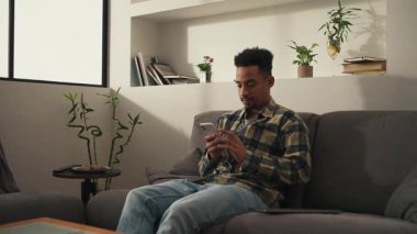 Gündelik kıyafetli Afrikalı adam modern oturma odasında kanepede oturuyor ve telefonda kayıyor. Yüksek kalite 4k görüntü