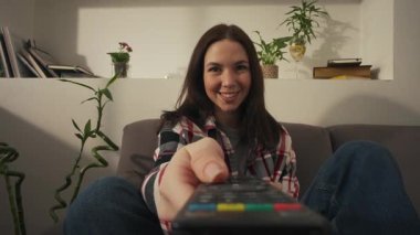 Ekose gömlekli genç esmer kadın kanepede oturuyor ve uzaktan kumandayla televizyon kanallarını değiştiriyor. Yüksek kalite 4k görüntü