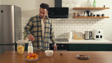 Yorgun Afrikalı Amerikalı bir adam mutfakta kahvaltı hazırlıyor, mısır gevreğine süt döküyor, uykulu bir sabah rutini. Yüksek kalite 4k görüntü