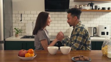 Mutfak masasında el ele tutuşan çok kültürlü bir çift. Birbirlerine bakıyorlar, gülümsüyorlar ve sıcak ve yumuşak bir sabah atmosferinde konuşuyorlar. Yüksek kalite 4k görüntü