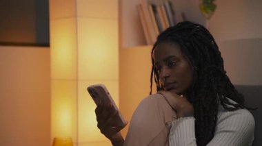 Akşamları kanepede oturan genç Afrikalı Amerikalı kadın, çenesini dizine dayayıp, akıllı telefonuna ciddi ciddi bakıyor. Yüksek kalite 4k görüntü