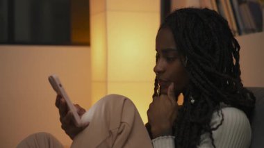 Genç bir Afro-Amerikalı kadının yan profil portresi, akıllı telefonuna odaklanmış bir ifadeyle bakıyor, saçlarını hafifçe kaydırırken ayarlıyor. Yüksek kalite 4k görüntü