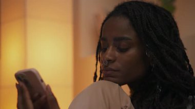 Akşamları kanepede oturan, çenesini dizlerinin üzerinde dinlendiren ve akıllı telefonuna odaklanmış bir ifadeyle bakan genç bir Afrikalı Amerikalı kadının portresi. Yüksek kalite 4k görüntü