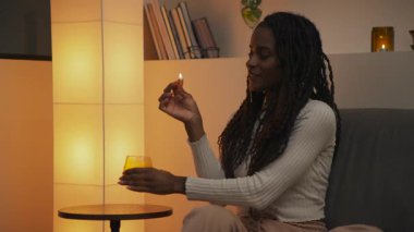 Genç Afrikalı Amerikalı kadın bir kibritle mum yakıyor ve onu bir masaya yerleştiriyor, sonra rahat bir şekilde oturuyor ve sıcak bir akşam ortamında aleve bakıyor. Yüksek kalite 4k görüntü