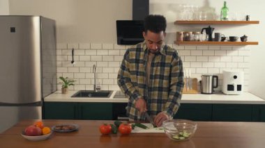 Sakin bir sabah sahnesinde, mutfakta salatalık doğrayan Afro-Amerikalı bir adam taze salata hazırlarken. Atmosfer huzurlu ve odaklanmış hissettiriyor. Yüksek kalite 4k görüntü