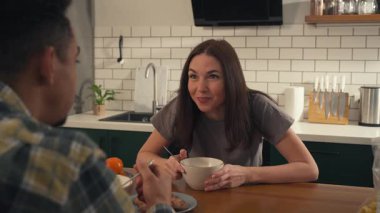 Genç bir çift sıcak bir sabahı paylaşıyor. El ele tutuşuyorlar, mısır gevreği yiyorlar ve güneşli bir mutfakta sohbet ediyorlar. Omzunun üstünden vurmak samimiyet ve samimi gerçekçilik ekler. Yüksek kalite 4k görüntü