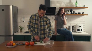 Afro-Amerikalı adam mutfakta tezgahta oturup telefonda konuşan kız arkadaşına gülümserken taze bir salata hazırlıyor. Yüksek kalite 4k görüntü