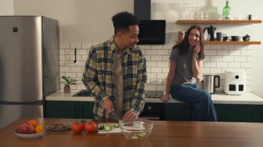 Afro-Amerikalı bir adam mutfakta salatalık dilimliyor ve telefonda sohbet eden gülümseyen kız arkadaşına bir ısırık veriyor. Aşk ve ışıkla dolu mutlu bir sabah sahnesi. Yüksek kalite 4k görüntü