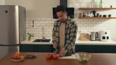 Afro-Amerikalı bir adam huzurlu bir sabahta mutfakta taze bir salata hazırlıyor. Kulaklık takıyor, gülümsüyor ve sebze doğrarken hafifçe dans ediyor. Yüksek kalite 4k görüntü