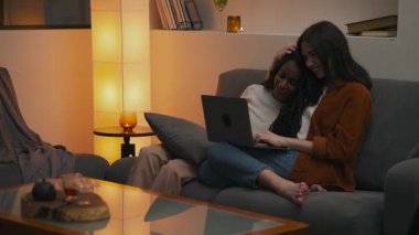 Sıcak ve rahat bir anda, çok ırklı lezbiyen bir çift, huzurlu bir yuva ortamında dizüstü bilgisayar kullanırken koltukta yan yana oturup, birbirlerine eşlik ediyorlar. Yüksek kalite 4k görüntü