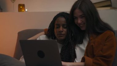 Çok kültürlü lezbiyen bir çiftin kanepede sarılıp, sohbet ettiği ve dizüstü bilgisayarda birlikte göz gezdirirken gülümsediği samimi bir akşam sahnesi. Yüksek kalite 4k görüntü