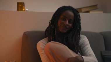 Rasta saçlı rahat bir Afro-Amerikalı kadın evinde huzurlu bir akşamın tadını çıkarıyor, kanepede oturuyor ve mum ışığına bakıyor. Yüksek kalite 4k görüntü