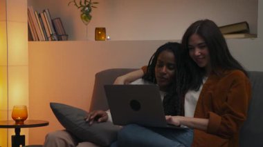Akşamları kanepede oturan çok ırklı lezbiyen çift dizüstü bilgisayarda birbirlerine bakıp gülümserken evlerinde sıcak ve samimi bir anı paylaşıyorlar. Yüksek kalite 4k görüntü