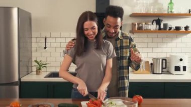 Çok kültürlü bir çift mutfakta neşeli bir sabahın tadını çıkarırken kamera geniş açıdan yakın plan çekime geçiyor. Kadın salatayı keserken erkek yakınlarda dans ediyor. Yüksek kalite 4k görüntü