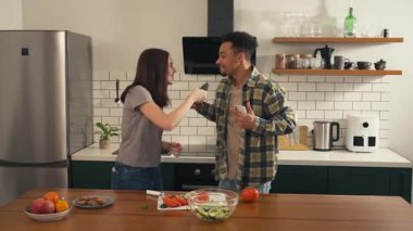Mutfakta neşeyle şarkı söyleyen ve dans eden çok kültürlü bir çift. Kadın mikrofon olarak salatalık kullanırken erkek telefonu tutuyor ve katılıyor. Yüksek kalite 4k görüntü