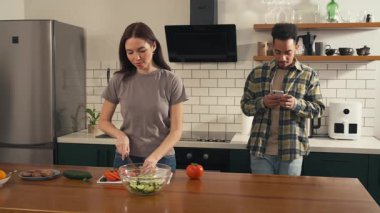 Kulaklıkları paylaştıktan sonra mutfakta dans eden çok kültürlü bir çift. Kadın salata hazırlarken erkek telefonunda müzik buluyor. Yüksek kalite 4k görüntü