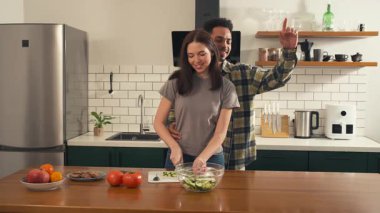 Çok kültürlü çiftler dans ediyor ve neşeli bir sabahta mutfakta taze salata yapıyorlar. Yüksek kalite 4k görüntü