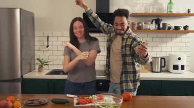 Çok kültürlü bir çift mutfakta neşeyle dans ediyor, müziğin tadını çıkarıyor ve taze salata hazırlarken duraklıyorlar. Yüksek kalite 4k görüntü