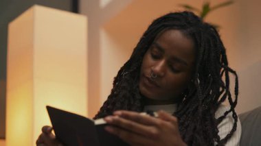 Afrika kökenli Amerikalı genç bir kadının yakın plan portresi kitap okuyor ve sayfaları yumuşak akşam ışığında çeviriyor, sakin ve rahat bir atmosfere daldırıyor. Yüksek kalite 4k görüntü