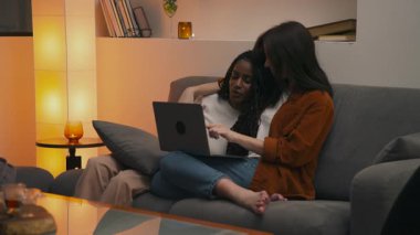 Çok ırklı lezbiyen çift, kanepede rahat bir akşamın tadını çıkarıyorlar. Birlikte dizüstü bilgisayar izlerken sarılıp gülümsüyorlar. Sıcak ve rahat bir ev ortamında samimi bir anı paylaşıyorlar. Yüksek kalite 4k görüntü